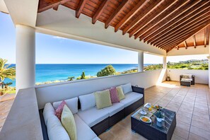 A covered outdoor seating area with a view of the sea, a sofa with cushions, a table with plates and glasses, and a wicker coffee table. - Oceanfront Luxury Villa with Heatable Infinity Pool & Breathtaking Ocean Views (Luz)