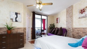 Habitación de hotel con cama, cómoda, ventilador de techo, planta en maceta y vista al balcón exterior.