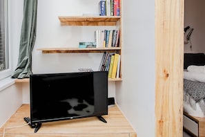 Une pièce avec un téléviseur posé sur un meuble, une bibliothèque remplie de livres, une fenêtre avec un rideau et un lit avec une couverture.