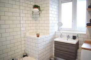 Une salle de bain avec des carreaux de céramique blancs rectangulaires, une petite fenêtre, un miroir et un lavabo doté d’un plan de travail en bois.