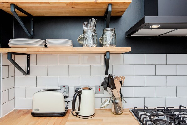A kitchen with a wooden shelf holding plates and jars, a toaster, a kettle, and a gas stove.