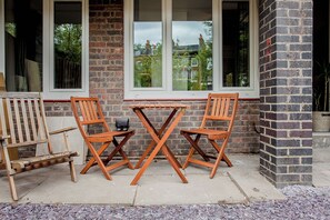 Une terrasse avec deux chaises en bois et une petite table, un mur en briques et des fenêtres donnant sur un quartier résidentiel.