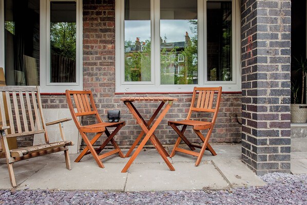 Eine Terrasse mit zwei Holzstühlen und einem kleinen Tisch, einer Ziegelwand und Fenstern, die auf ein Wohngebiet blicken.