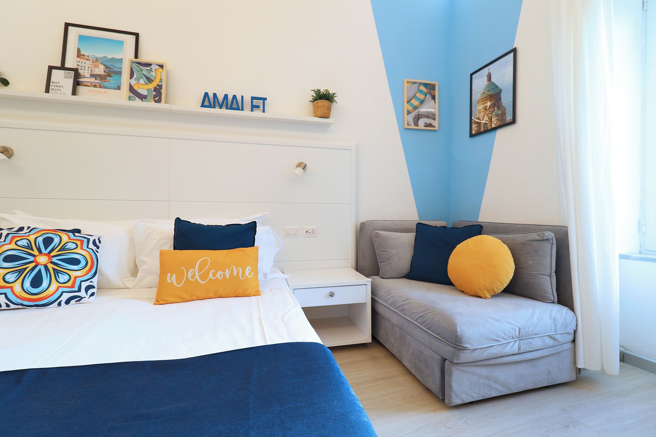 A modern bedroom with a bed, a grey sofa, and a nightstand. There are decorative pillows and a welcome sign on the bed.
