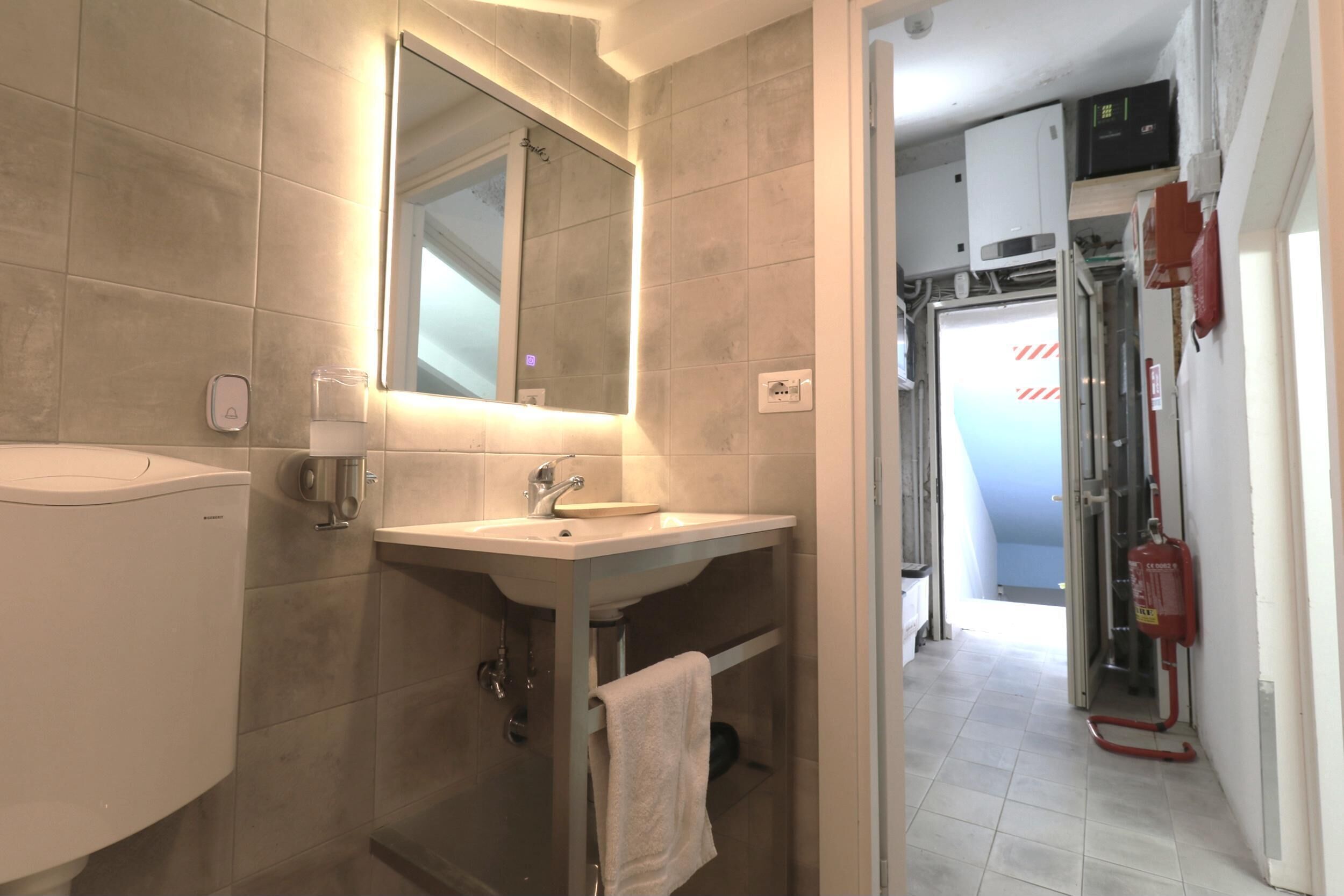 A modern bathroom with a sink, mirror, and toilet. There is a towel rack and a fire extinguisher visible.