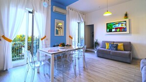 Un salon moderne avec un coin repas, comprenant un canapé, des chaises transparentes, une table et un téléviseur fixé au mur.