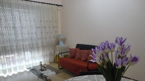 Ein Wohnzimmer mit einem roten Sofa, einem kleinen runden Tisch mit Spitze und einer Vase mit lila Blumen.