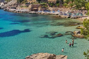 A coastal beach with clear turquoise water, rocky shorelines, and sun loungers.