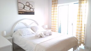 Un lit bien fait, avec du linge de lit et des serviettes blancs, une table de chevet blanche avec une lampe, et un tableau accroché au-dessus du lit.