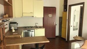 A compact kitchen with a dishwasher, stove, and sink. There is a wooden table and a black chair. A brown door leads to another room. - VerdeCielo in Marina di Massa, Eliana's cottage  (Marina di Massa)