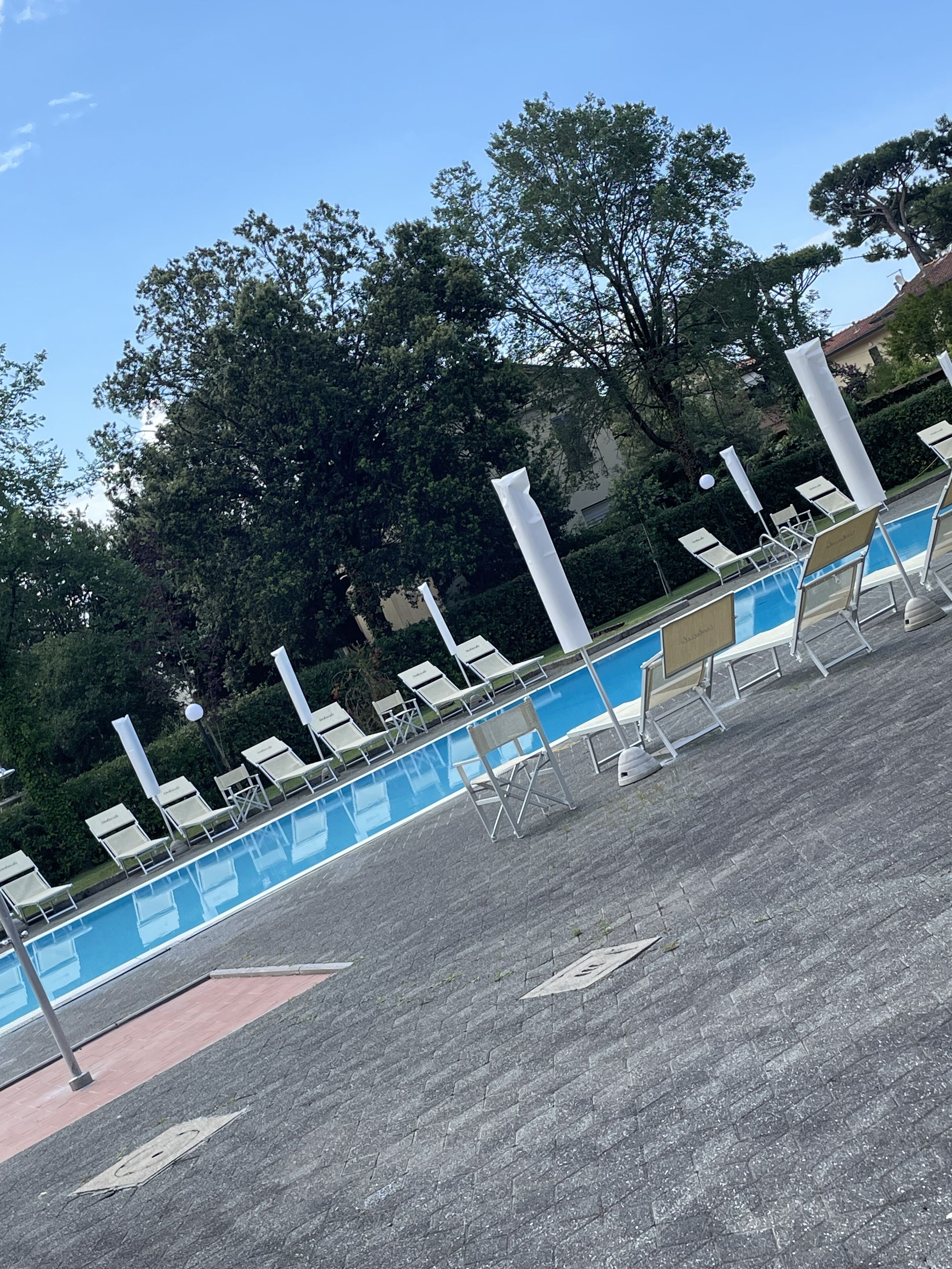 A poolside area with lounge chairs and umbrellas, surrounded by trees and a building in the background.