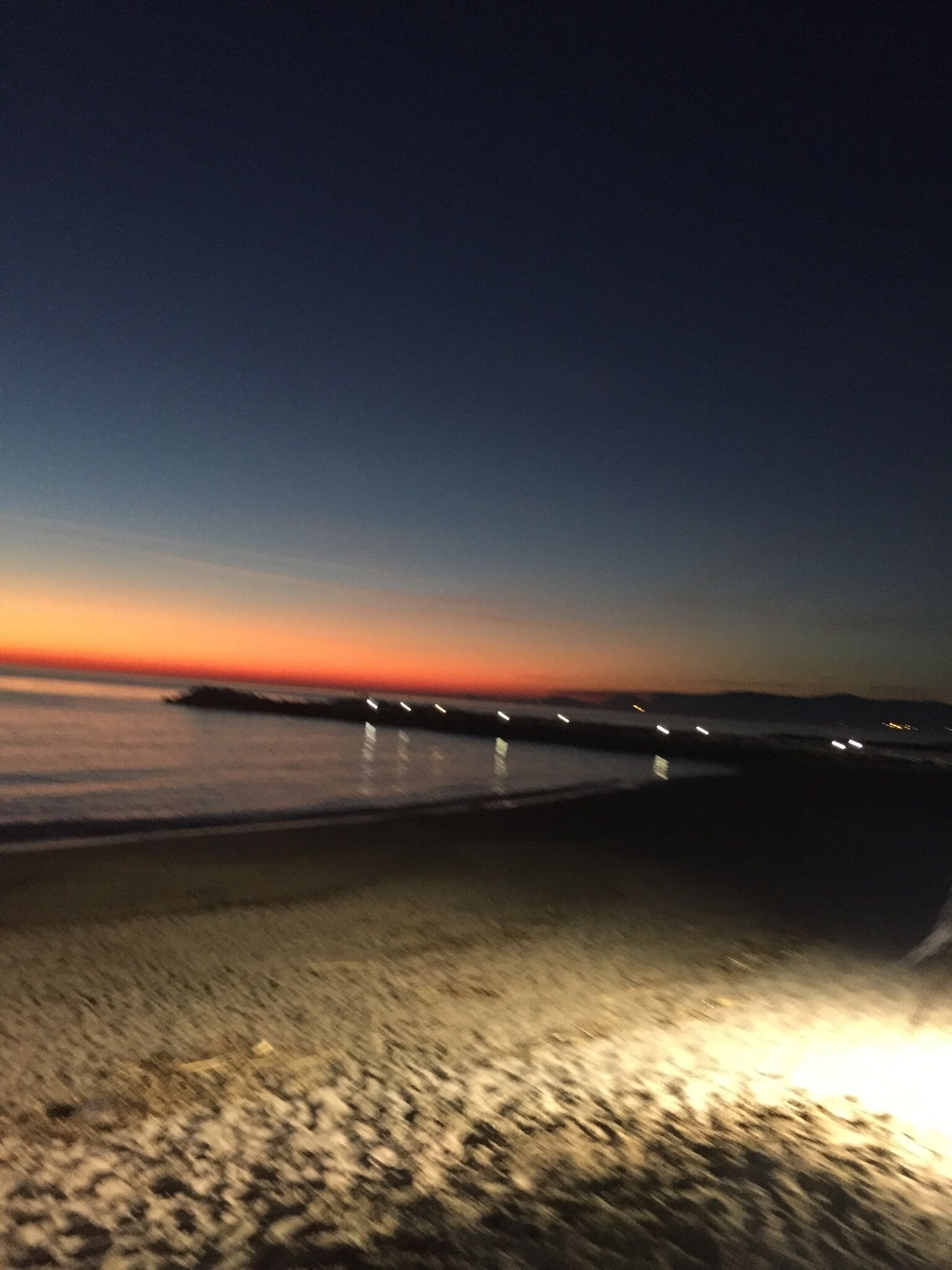Ein Strand im Abenddämmerung mit ruhiger See, einer sich ins Wasser erstreckenden Mole und einem sandigen Ufer.
