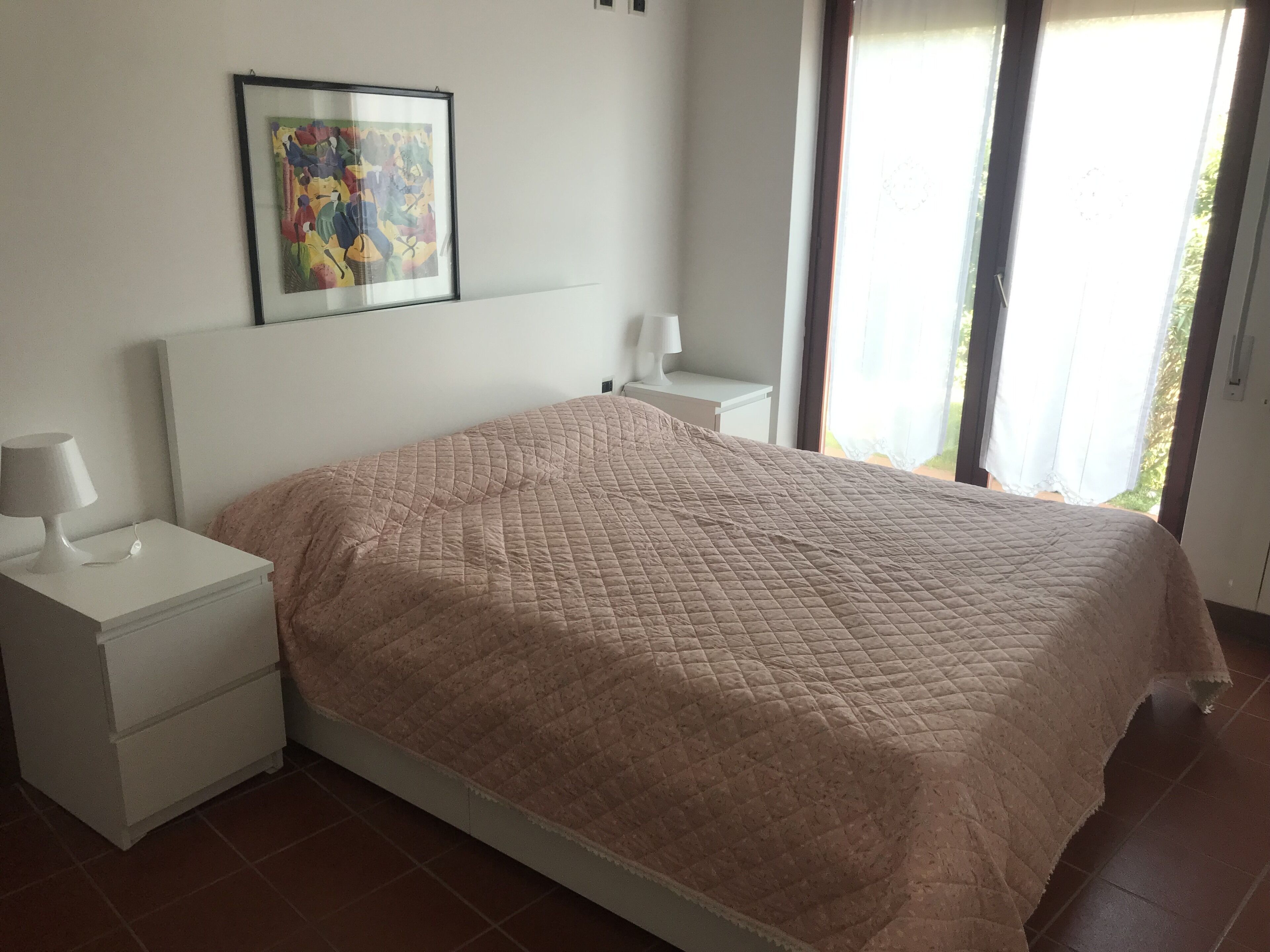 A bedroom with a bed, a nightstand, a bedside table, a framed picture on the wall, and a window with curtains.