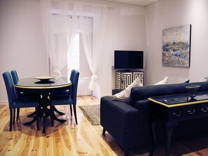Una sala de estar moderna con una mesa de comedor redonda, sillas azules, un sofá oscuro, un televisor de pantalla plana y un cuadro decorativo en la pared.