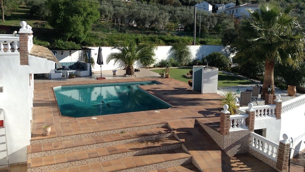 A pool area with steps leading down to the water, surrounded by a white wall and a garden with palm trees and a lawn. - Kayenne 2 ,2 bedroom self catering apartment (Iznajar)