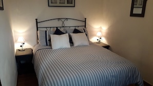 A neatly made bed with striped bedding, two bedside tables with lamps, and framed pictures on the walls. - Kayenne 2 ,2 bedroom self catering apartment (Iznajar)