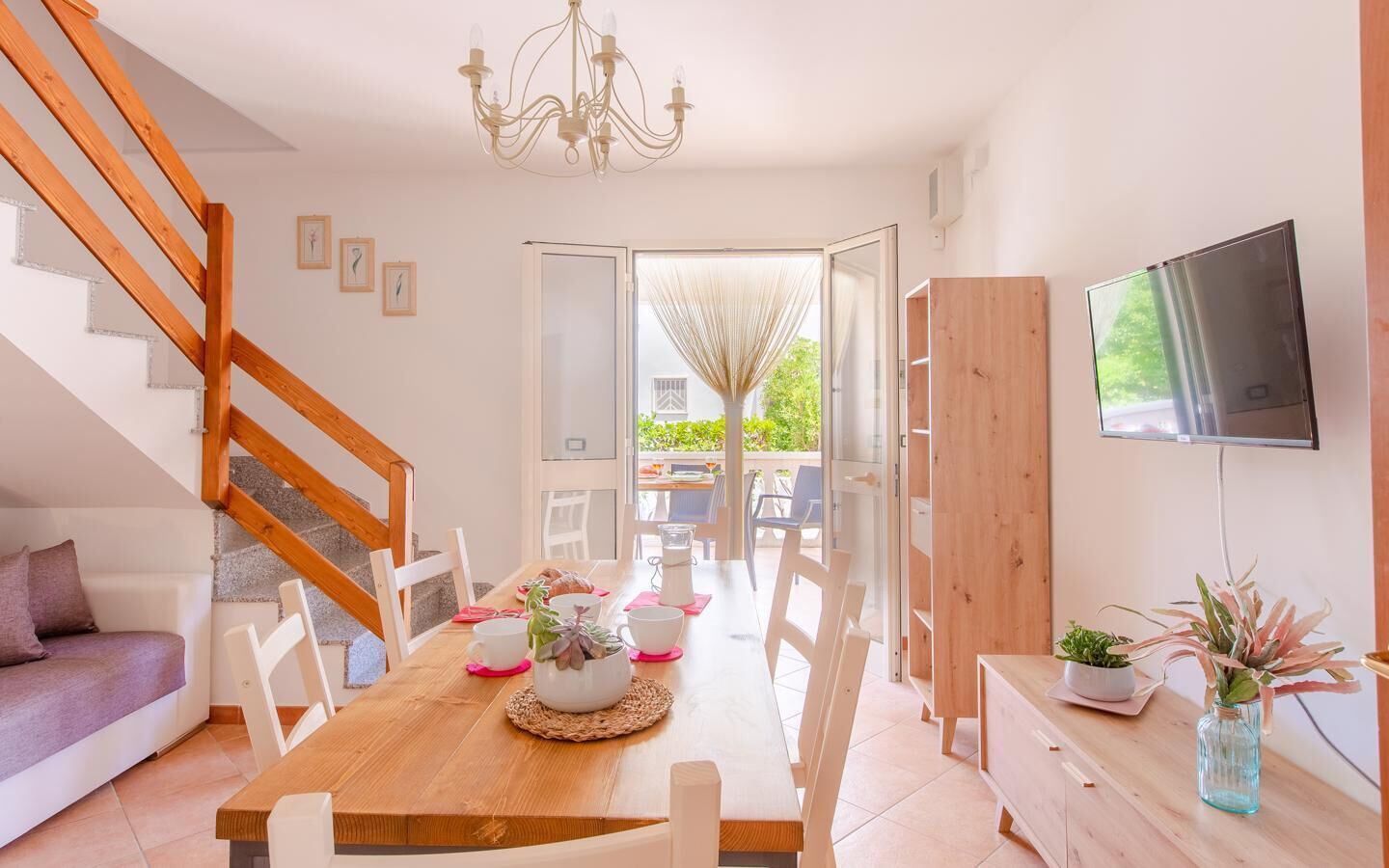 Een moderne eetruimte met een houten tafel, witte stoelen en een kroonluchter. Er is een televisie aan de muur bevestigd en een trap die naar een andere kamer leidt.