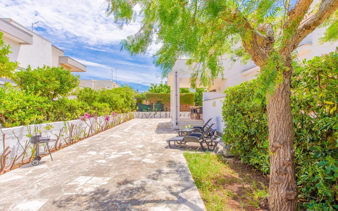 Une terrasse dallée, aménagée avec du mobilier d’extérieur, entourée d’une végétation luxuriante et d’un arbre.