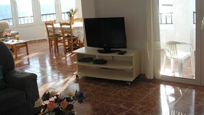 A living room with a television on a stand, a dining area with a table and chairs, and a balcony with a view. - House in Calan Morell with garden, private pool and BBQ 300 m from the beach (ciudadela de menorca cALAN morell)
