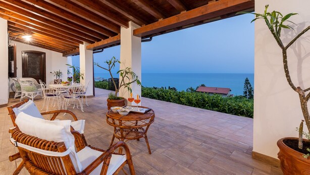 Un patio al aire libre con mobiliario de mimbre, una mesa con bebidas y aperitivos, y vistas al mar.