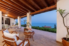 Une terrasse extérieure avec des meubles en osier, une table dressée avec des boissons et des amuse-gueules, et une vue sur la mer.