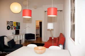 A modern living room with a white dining table, red and black sofas, and a wooden bowl centerpiece. There are two pendant lights, a white floor lamp, and a bookshelf with a television. A colorful abstract painting is on the wall.