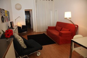 A compact living space with a red sofa, a black armchair, a small table, and a lamp. A white curtain and a door are visible in the background.