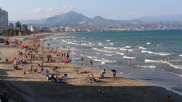 Una platja amb gent que es bronzeja i nedegui, edificis al fons i muntanyes a la distància.