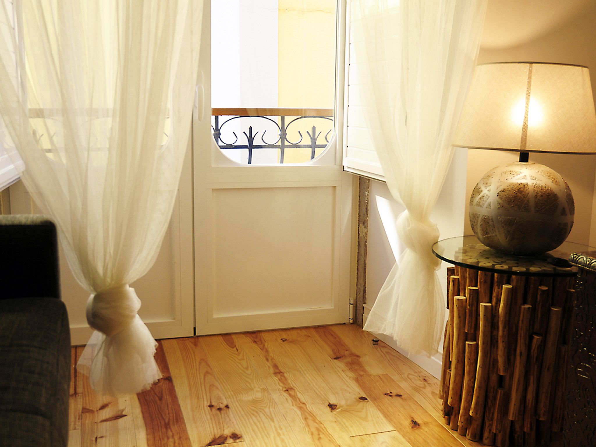 Una habitación con piso de madera, una puerta blanca con reja metálica, una mesa de vidrio con lámpara y cortinas de tela ligera.