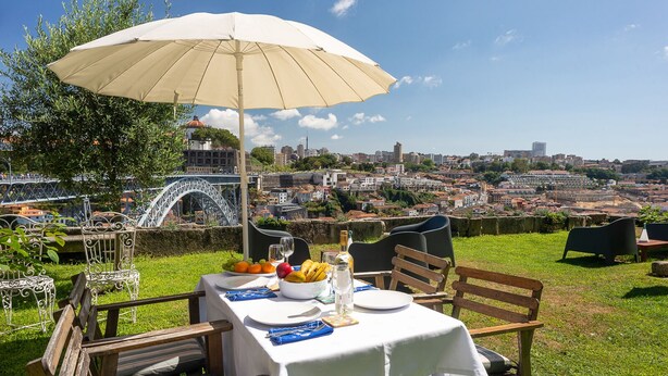 Un área de comedor al aire libre con una mesa para dos, un paraguas grande y una vista panorámica de la ciudad de fondo.