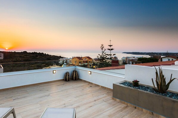 A rooftop terrace with a view of the sunset over the sea, wooden flooring, and a few potted plants. - Luxury Villa Amelia (Chania)