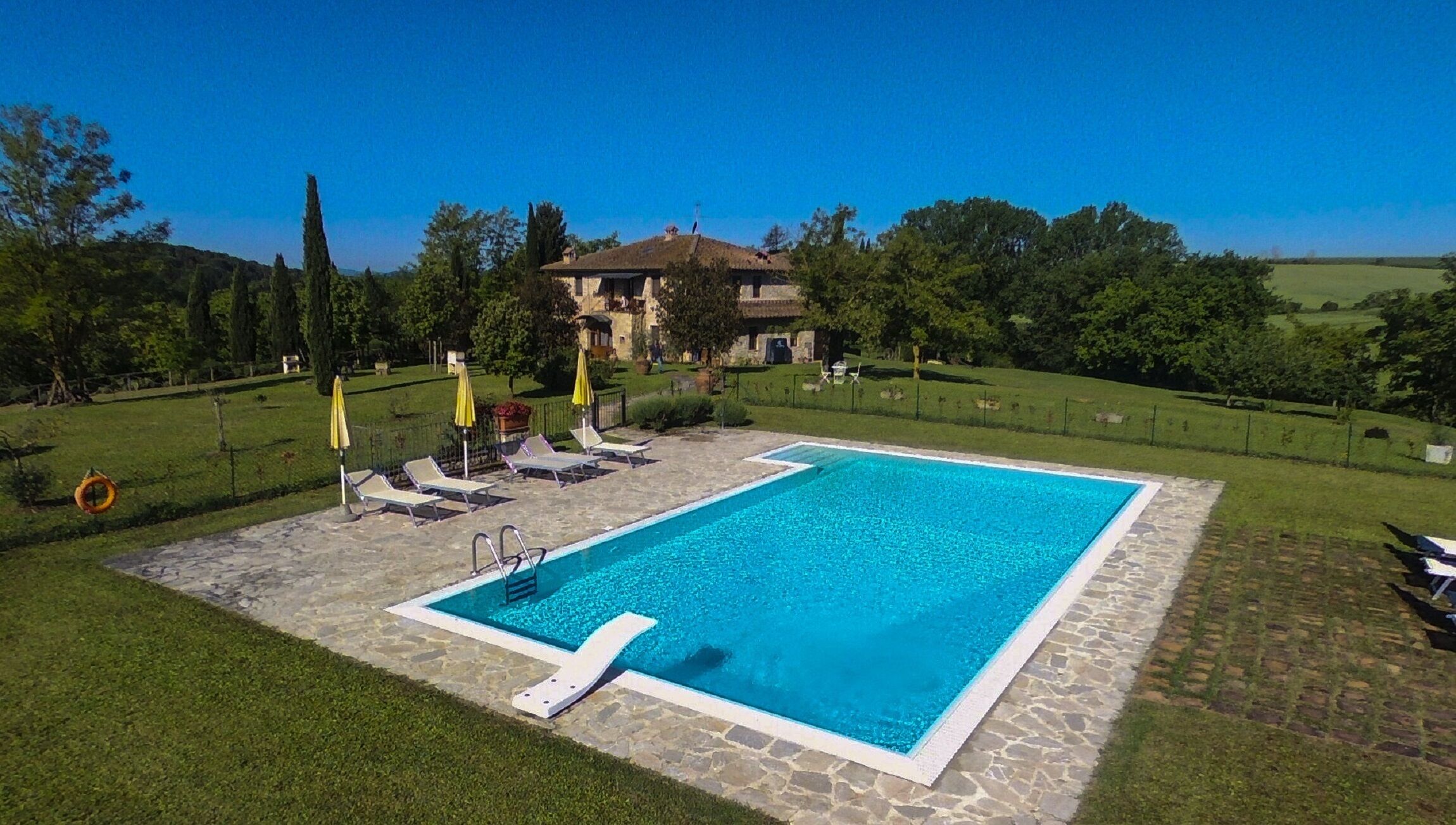 Ein rechteckiger Swimmingpool mit Steinrand, umgeben von einer Rasenfläche mit Liegestühlen und Sonnenschirmen.