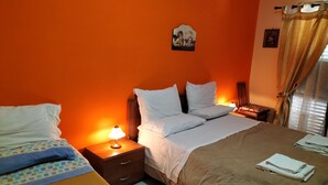 Ein Hotelzimmer mit oranger Wand, einem Bett mit weißer Bettwäsche, einem Nachttisch mit einer Lampe und einem Fenster mit Vorhängen.