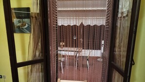 Ein Zimmer mit einem hölzernen Stuhl vor einem blickdichten Vorhang mit Muster. Im Hintergrund stehen ein Tisch und ein Stuhl.