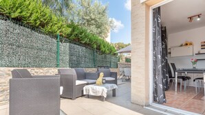 Une terrasse extérieure avec des meubles en osier, un chien et une vue sur la cuisine à travers une porte vitrée.