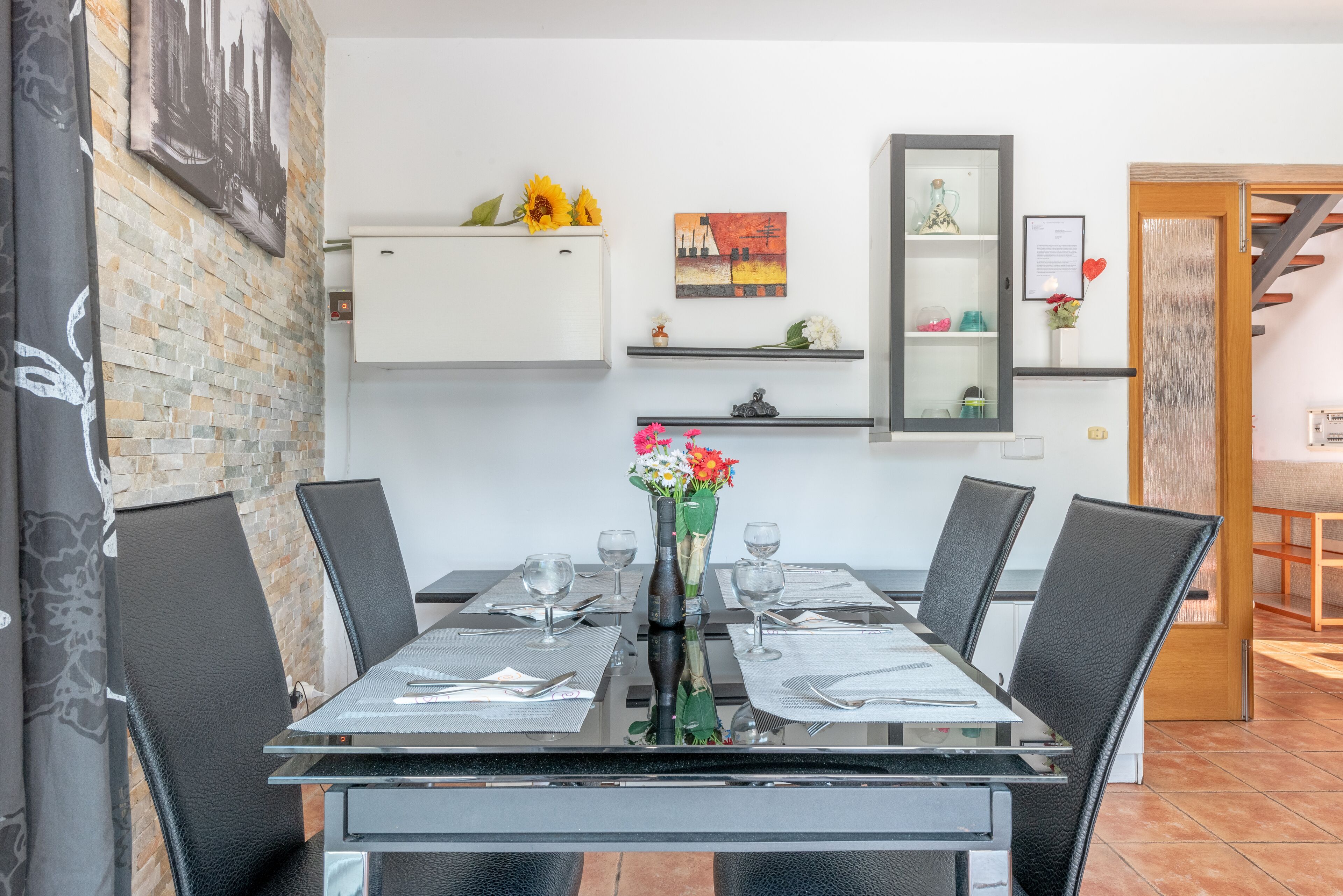 Un comedor con mesa de vidrio, sillas negras y un jarrón con flores. Se observan estantes empotrados con objetos decorativos y un cuadro.