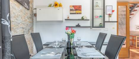 Un comedor con mesa de vidrio, sillas negras y un jarrón con flores. Se observan estantes empotrados con objetos decorativos y un cuadro.