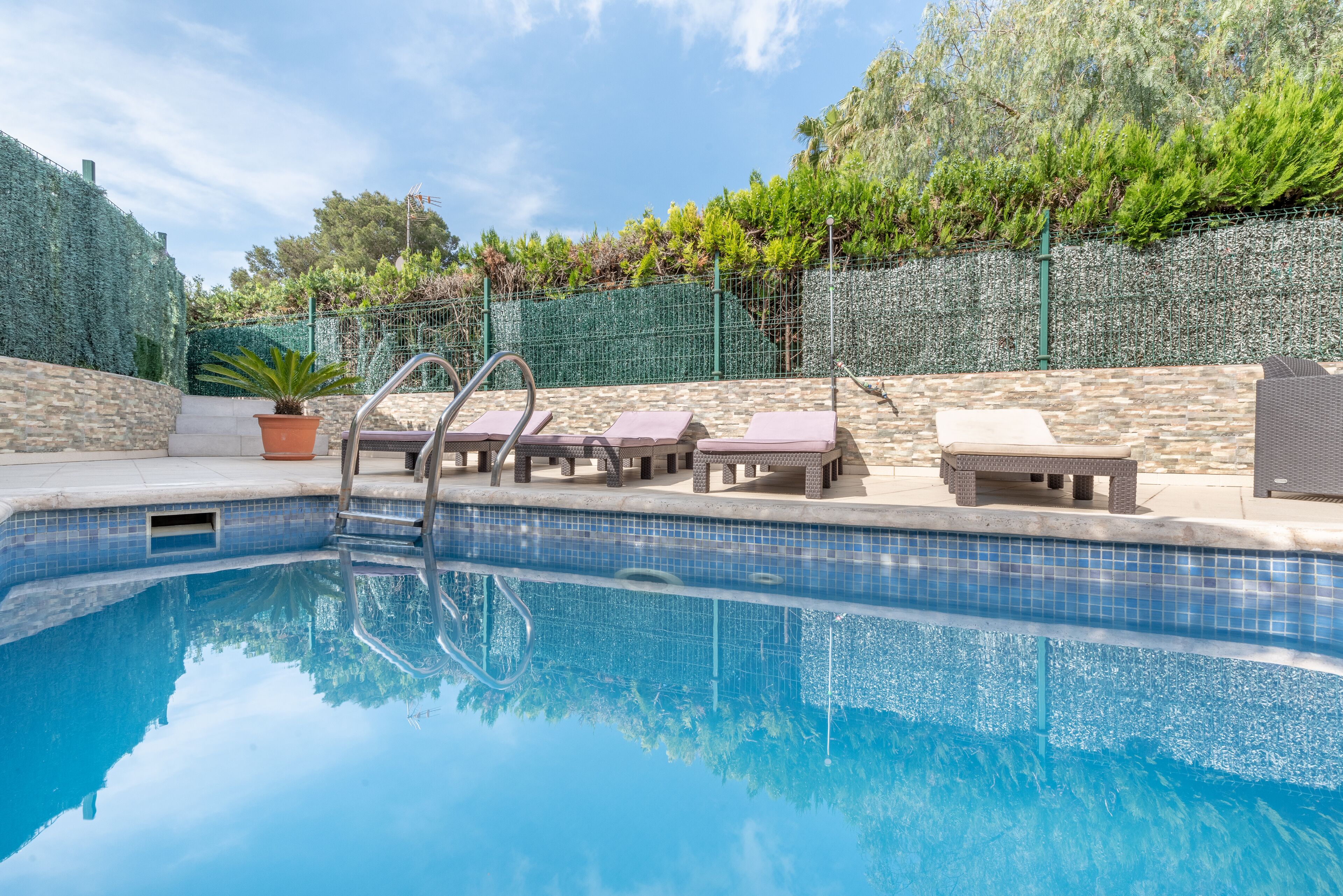 A swimming pool with lounge chairs and a small table, surrounded by a stone wall and a green fence.