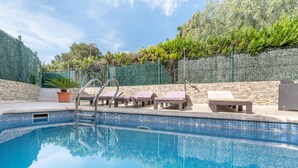 Une piscine entourée de chaises longues et d’une petite table, le tout délimité par un mur de pierre et une clôture verte.