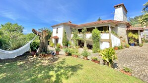 Ein zweistöckiges Steinhaus mit Ziegeldach, umgeben von einem gepflegten Garten mit verschiedenen Kübelpflanzen und einer Hängematte.