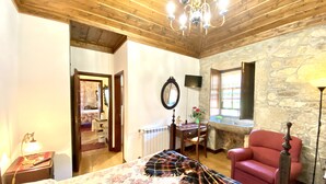 Ein Zimmer mit Holzbalkendecke, einem Bett mit floralem Bettzeug, einem roten Sessel, einer Steinwand und einer Pendelleuchte.