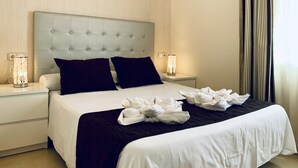 Hotellihuone, jossa on napitettu pääty, kaksi yöpöytää, valkoiset vuoteeksi, tummat tyynyt ja koristeellinen valkoinen pyyheasetelma.