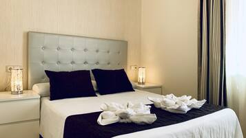 Habitación de hotel con tablero de cabecera capitonado, dos mesitas de noche, ropa de cama blanca, almohadas oscuras y una decoración de toallas blancas.