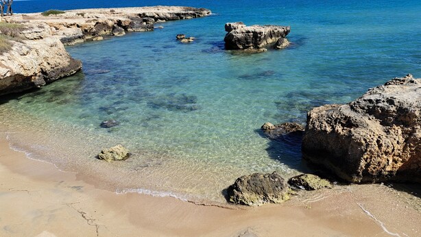 A coastal beach with clear water, rocky formations, and sandy shore.