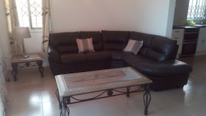 A living room with a dark brown leather sectional sofa, a glass-top coffee table, a small wooden side table, and a window with curtains. - Chic One bedroom serviced  apartment with  pool- fully self contained  V6 (Accra)