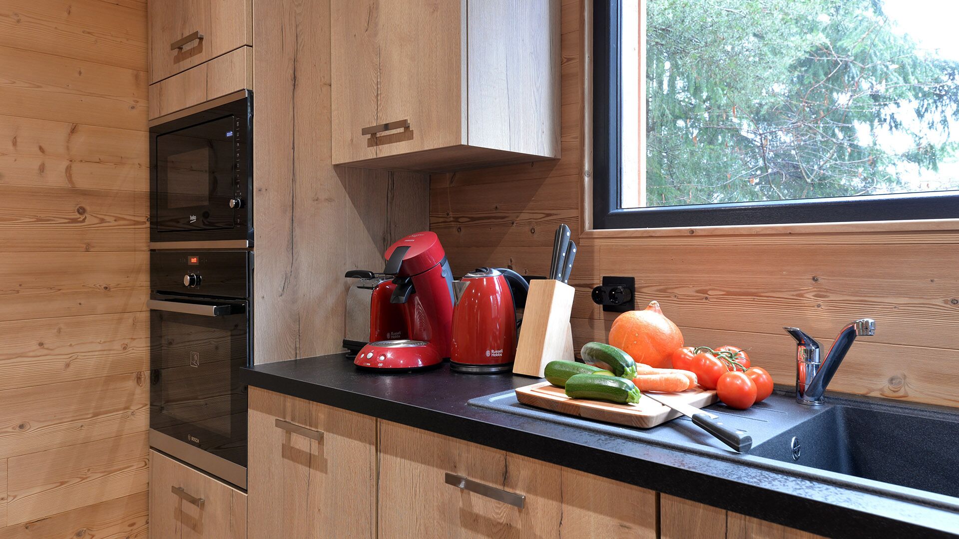 Una cocina moderna con armarios de madera, horno empotrado, cafetera roja y verduras frescas en la encimera.
