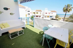 A balcony with a white sofa, a small table, and chairs, overlooking a beach and colorful buildings.