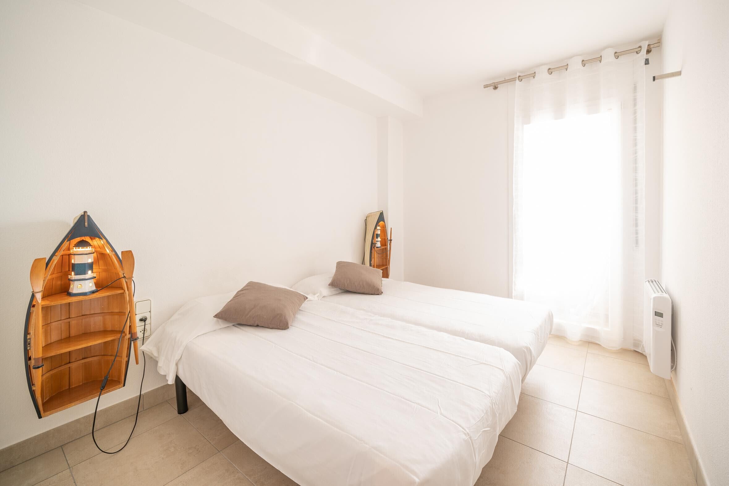 Une chambre propre et bien éclairée, avec deux lits, une étagère en bois sur laquelle se trouve une lampe, et une fenêtre ornée de rideaux légers.