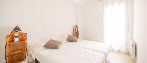 Une chambre propre et bien éclairée, avec deux lits, une étagère en bois sur laquelle se trouve une lampe, et une fenêtre ornée de rideaux légers.