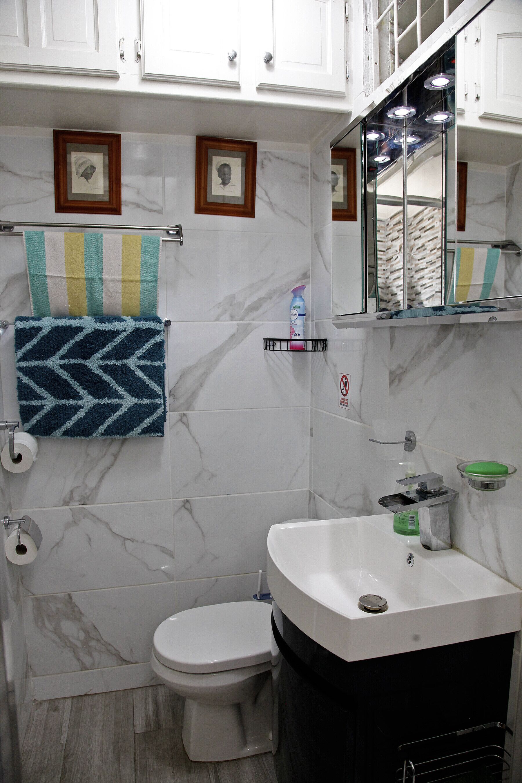 Baño con inodoro, lavamanos y paredes de mármol. Toallas cuelgan de un perchero y se observa una zona de ducha al fondo.